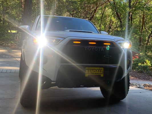 White 5th gen Toyota 4Runner with amber grille lights and headlights on among trees
